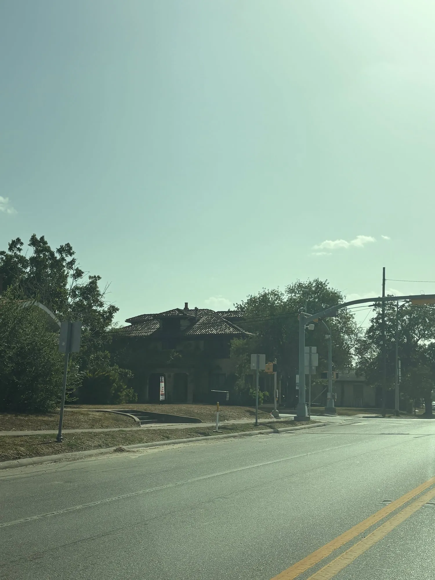 Weathered two story house with a clay tile roof and arched entry, half hidden by trees, beside a traffic signal and an empty street
