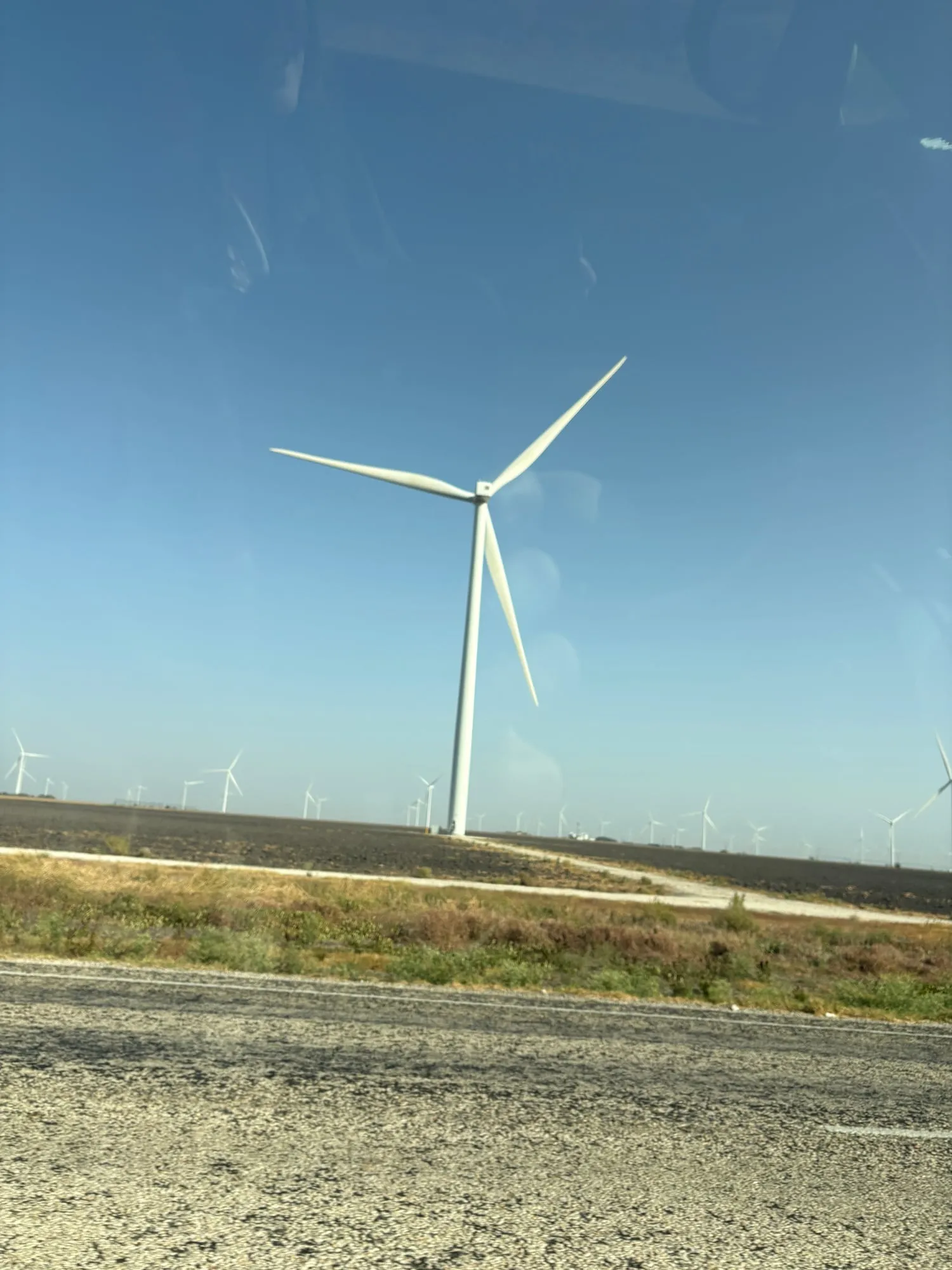 White wind turbine towering over a dark plowed field, with dozens of smaller turbines lined up along the horizon under a clear blue sky