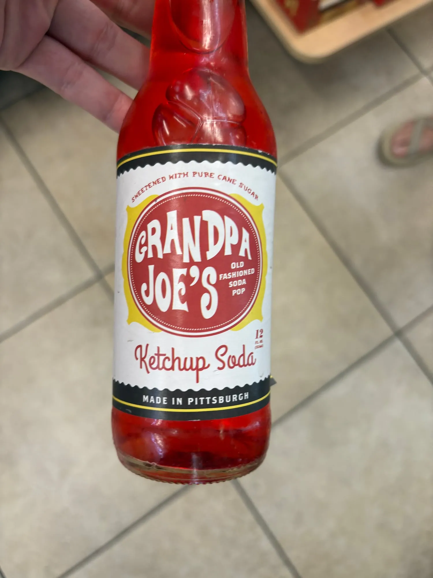 Hand holding a glass bottle of bright red Grandpa Joe's Ketchup Soda over a tile floor
