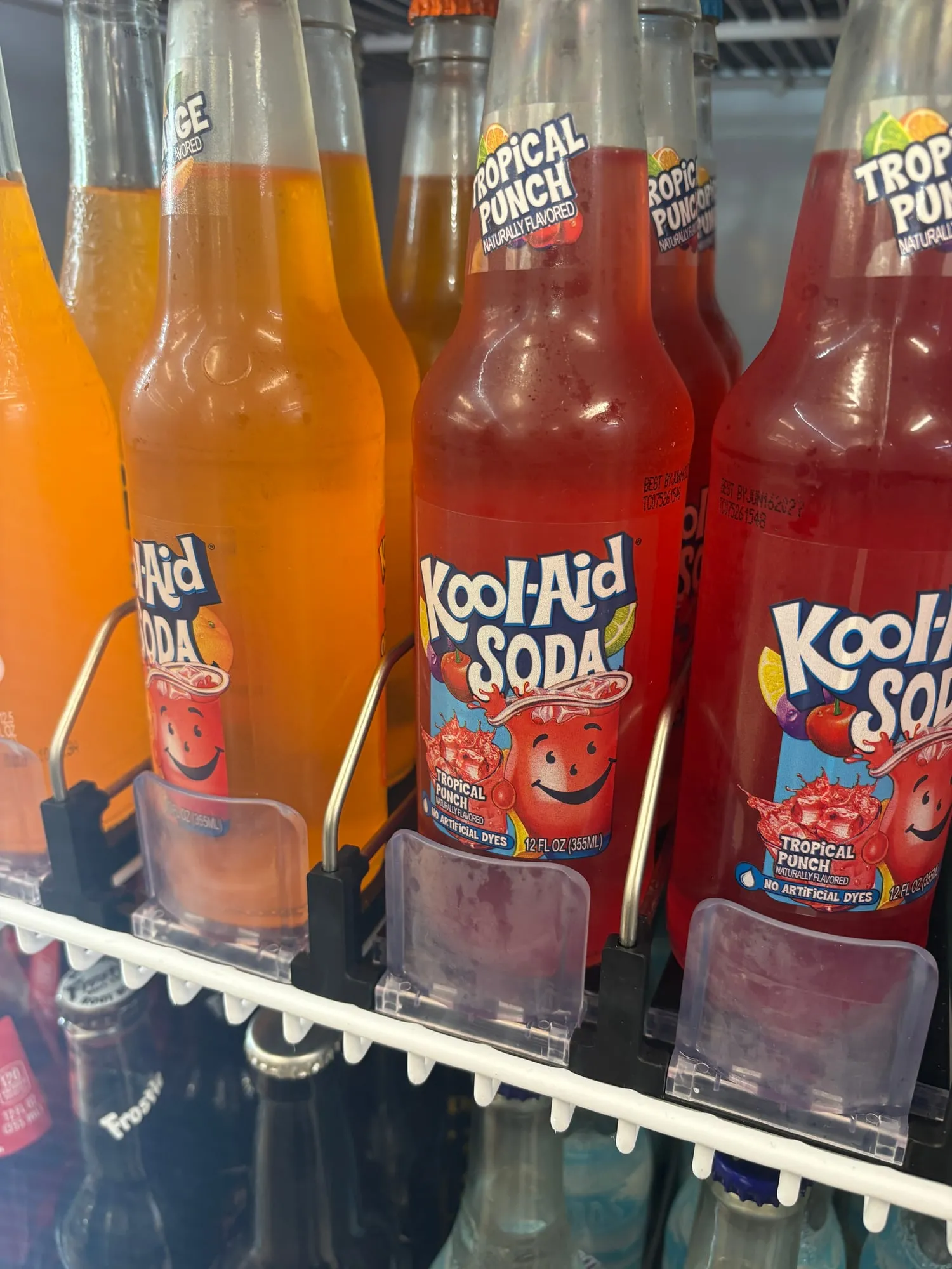 Cooler shelf of glass soda bottles, two red Kool-Aid Soda Tropical Punch bottles in front and orange ones behind