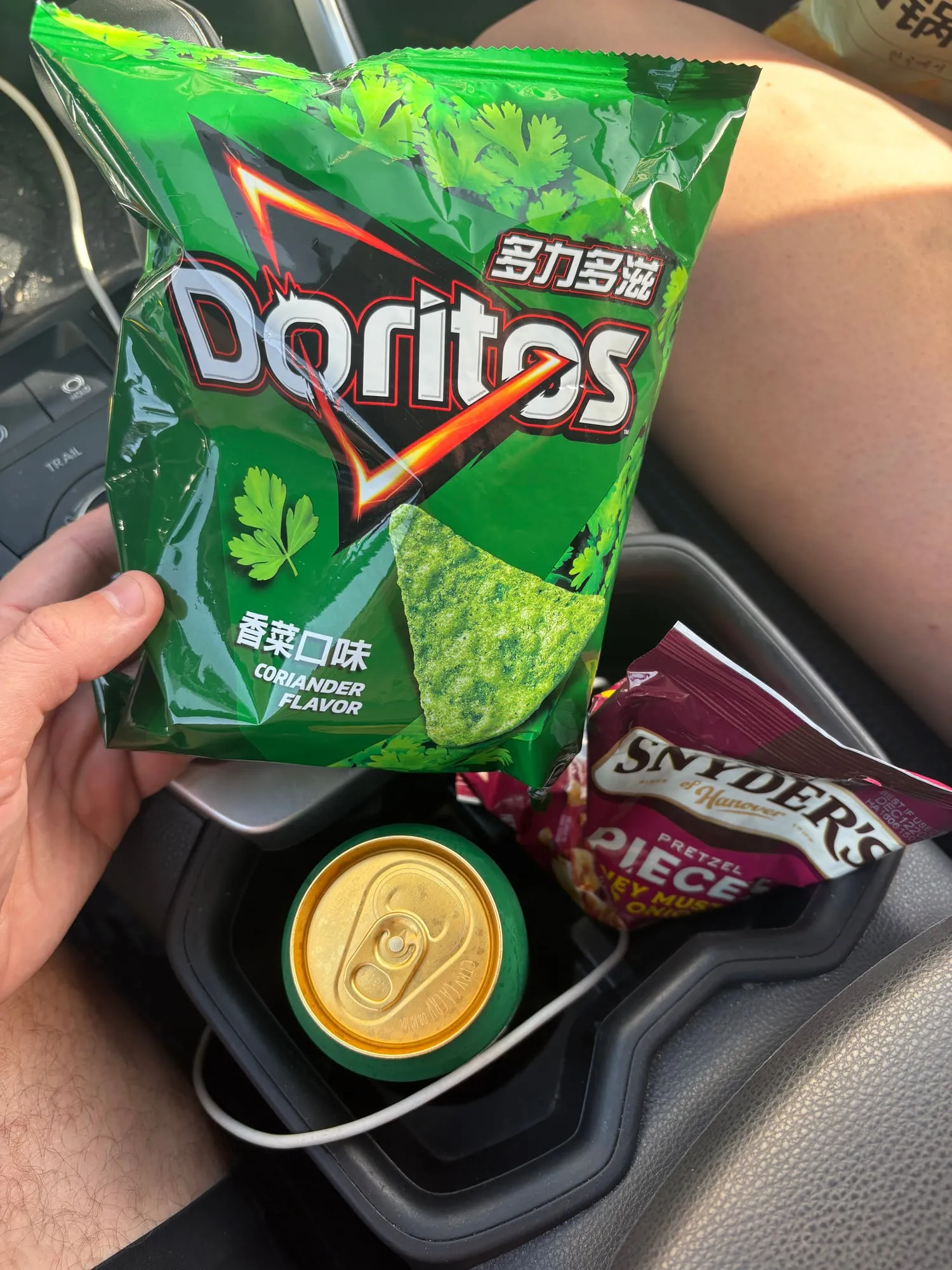 Green bag of coriander flavor Doritos held over a car console with a green can and a Snyder's pretzel pieces bag in the cupholder