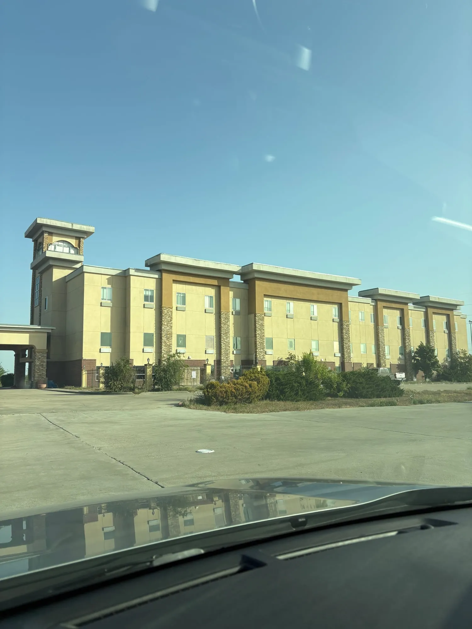 Three story tan hotel with boarded and blanked out windows behind a fence, empty concrete lot in front