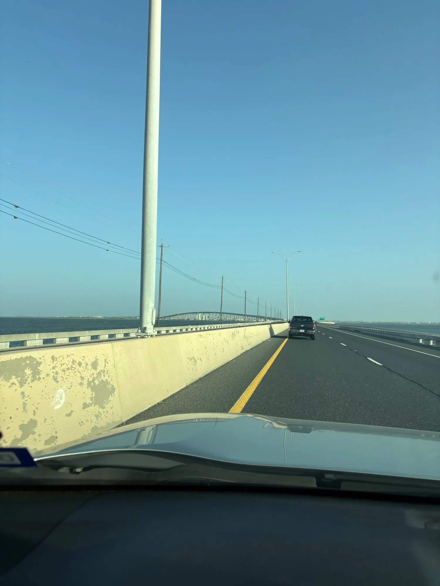View through a windshield of a divided causeway crossing open water, with the arch of the bridge ahead and a pickup truck in the next lane