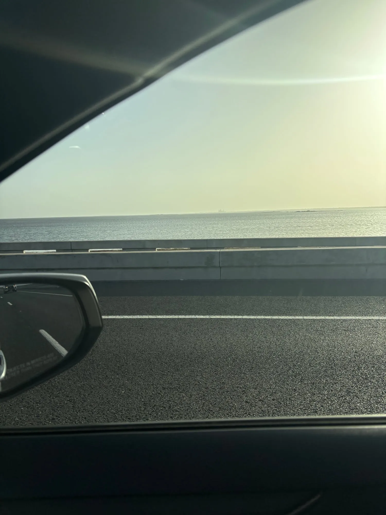 Flat hazy water of the Laguna Madre seen over a concrete barrier from the passenger window, low land on the far horizon