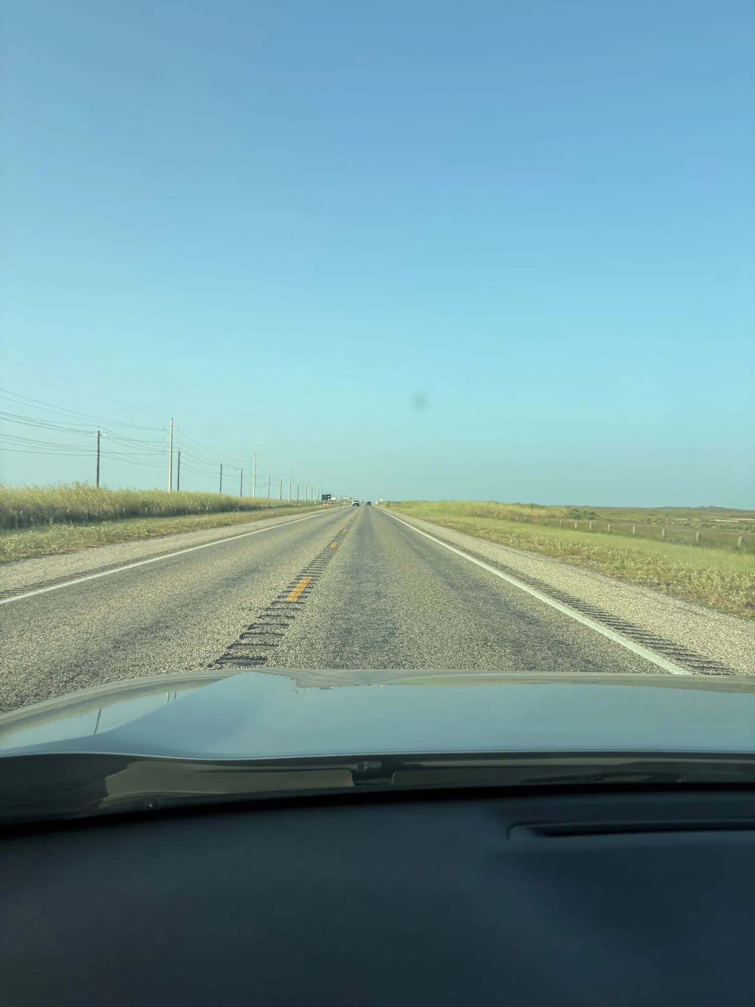Straight two lane road running to a flat horizon, tall grass and power lines on the left, low dunes on the right