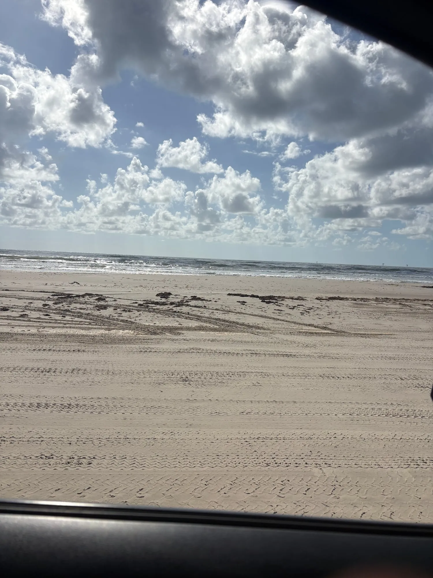 Wide sandy beach crisscrossed with tire tracks seen through a car window, the Gulf glittering beyond under a sky full of puffy clouds