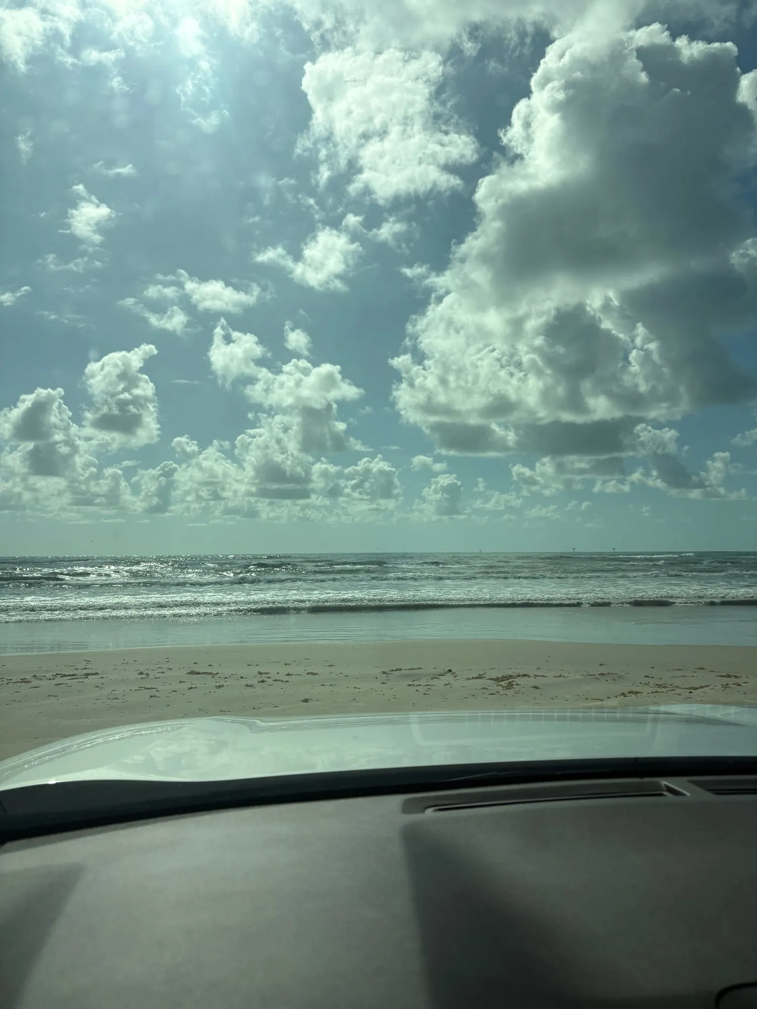 Gulf surf rolling onto a flat sandy beach seen over the hood of a car, with big white clouds stacked up against a blue sky
