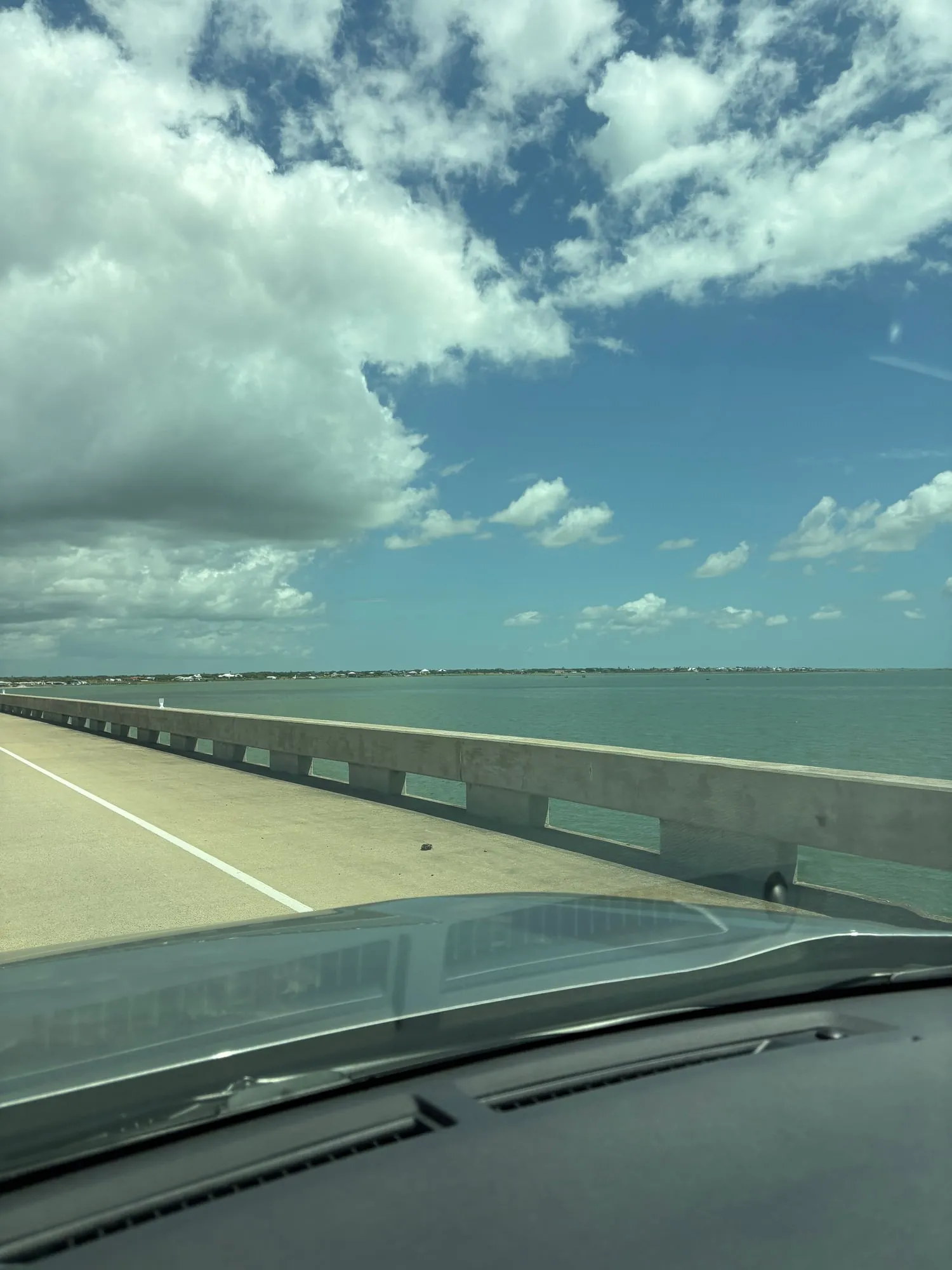 Green bay water beyond a low concrete bridge railing seen from a moving car, a thin strip of shoreline on the horizon under puffy clouds