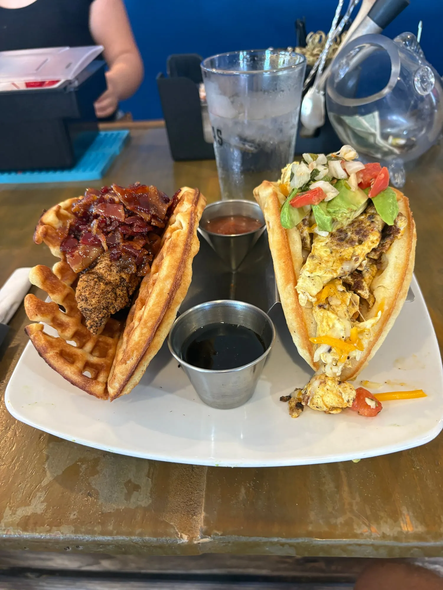 Two waffle tacos on a white plate, one stuffed with fried chicken and candied bacon, the other with eggs, cheese, avocado, and pico, with cups of syrup and salsa between them