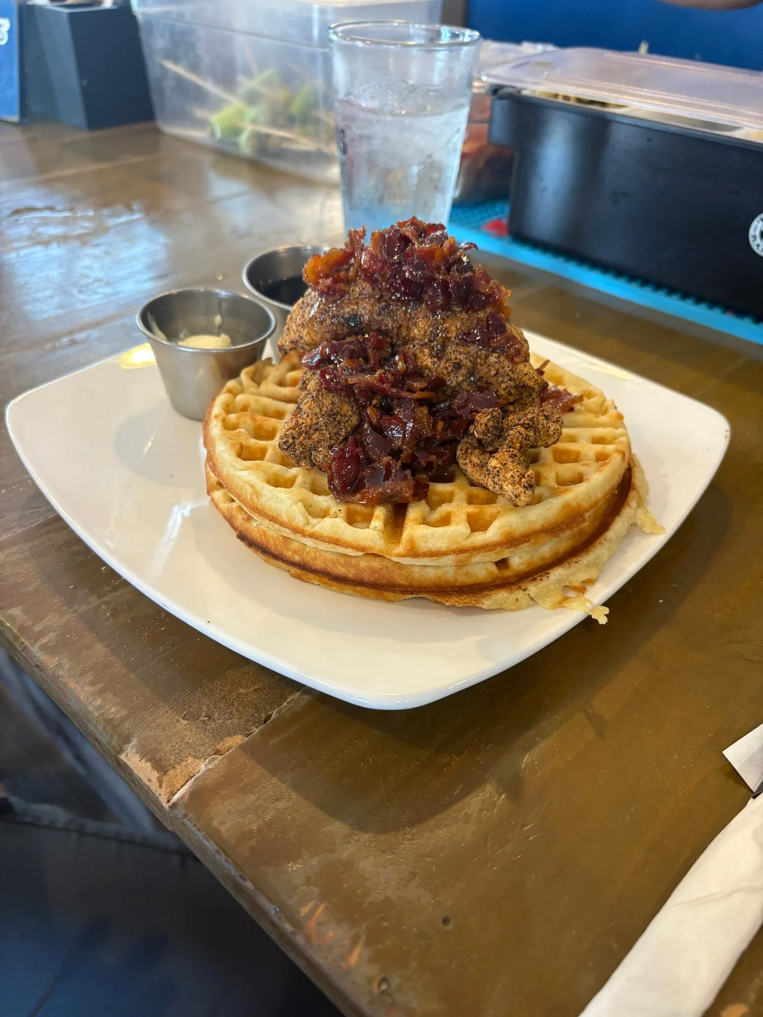 Stack of waffles topped with peppery fried chicken and a pile of candied bacon, with cups of butter and syrup on the side