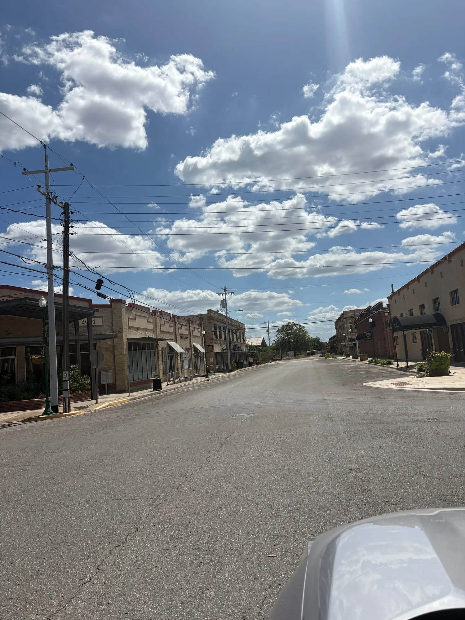 Empty small town street lined with low brick storefronts and utility poles under a bright sky full of clouds