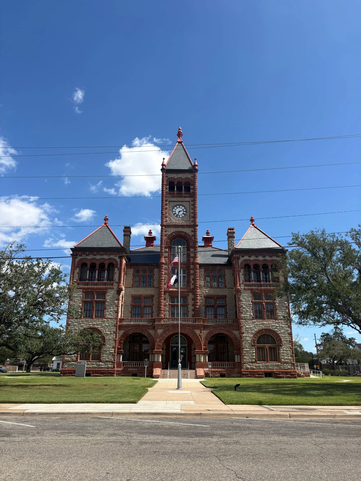 Three story stone courthouse with red sandstone arches, corner towers, and a tall central clock tower, flags flying out front under a deep blue sky