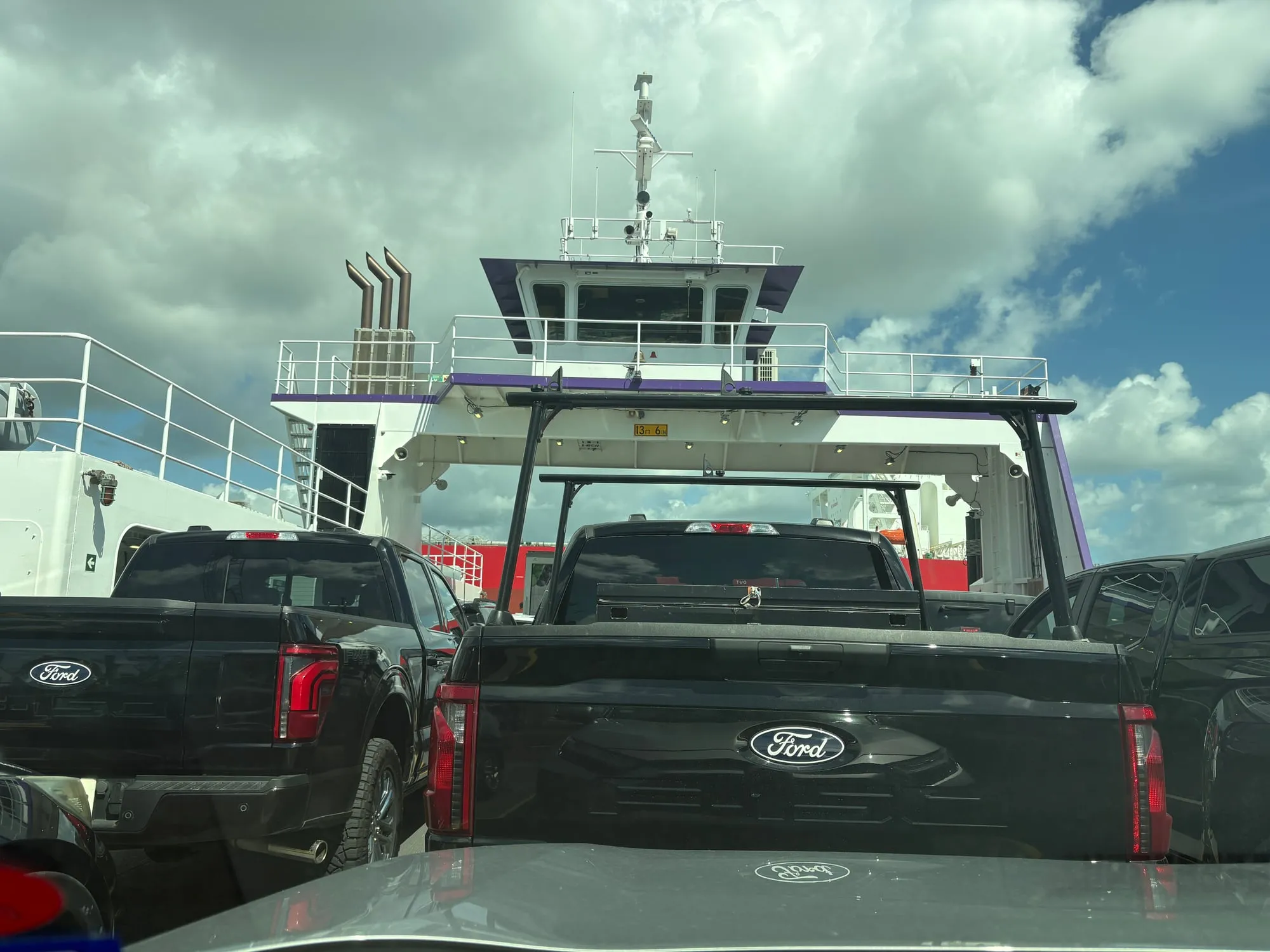 Wide view from the ferry deck of the pilothouse and twin exhaust stacks, with the red hull of a large ship passing behind the pickups