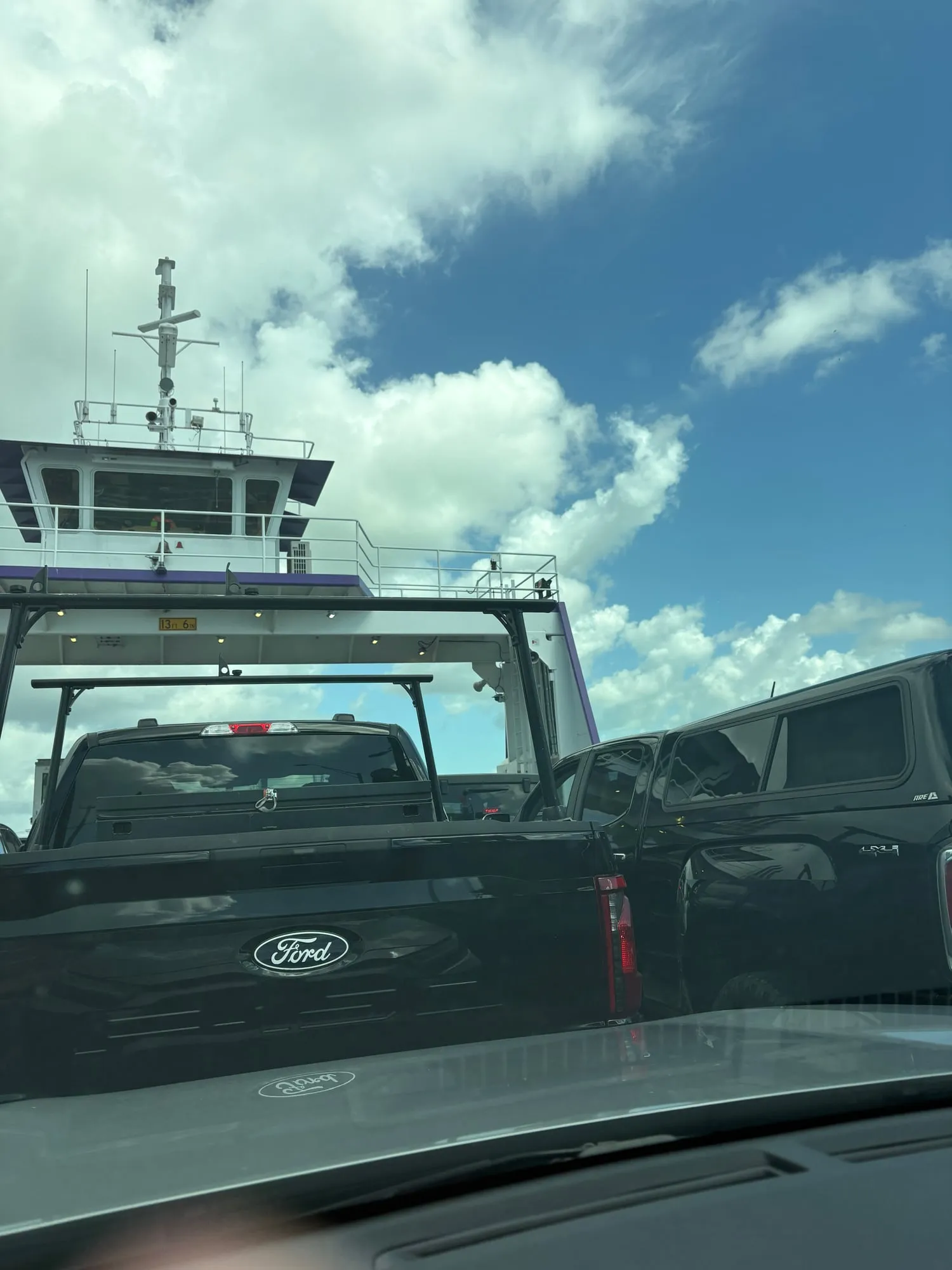 Ferry pilothouse with purple trim against a deep blue sky, seen over the ladder rack of the pickup parked in front