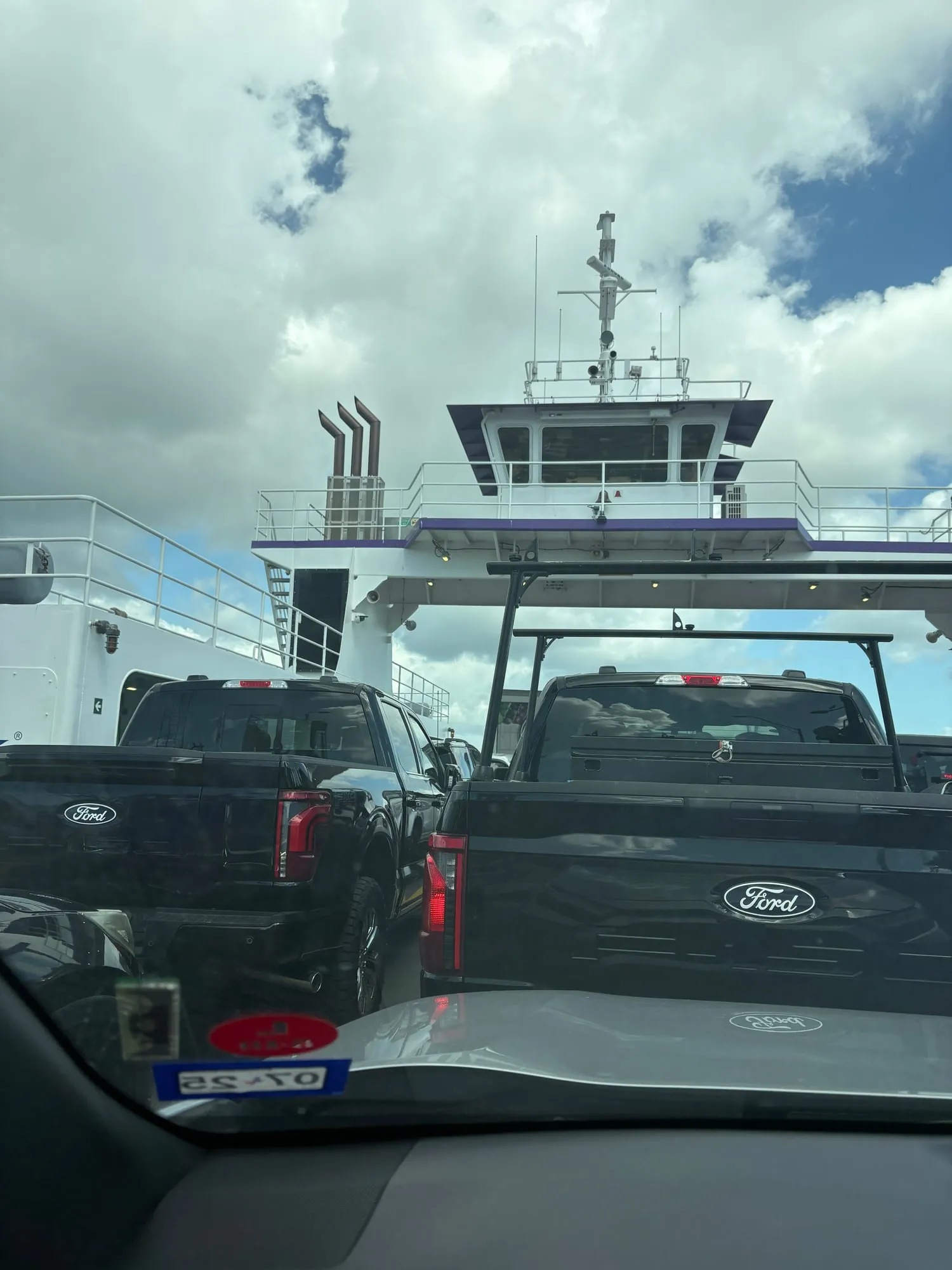 View through a windshield from the deck of a car ferry, two black pickups parked ahead and the white pilothouse rising above them