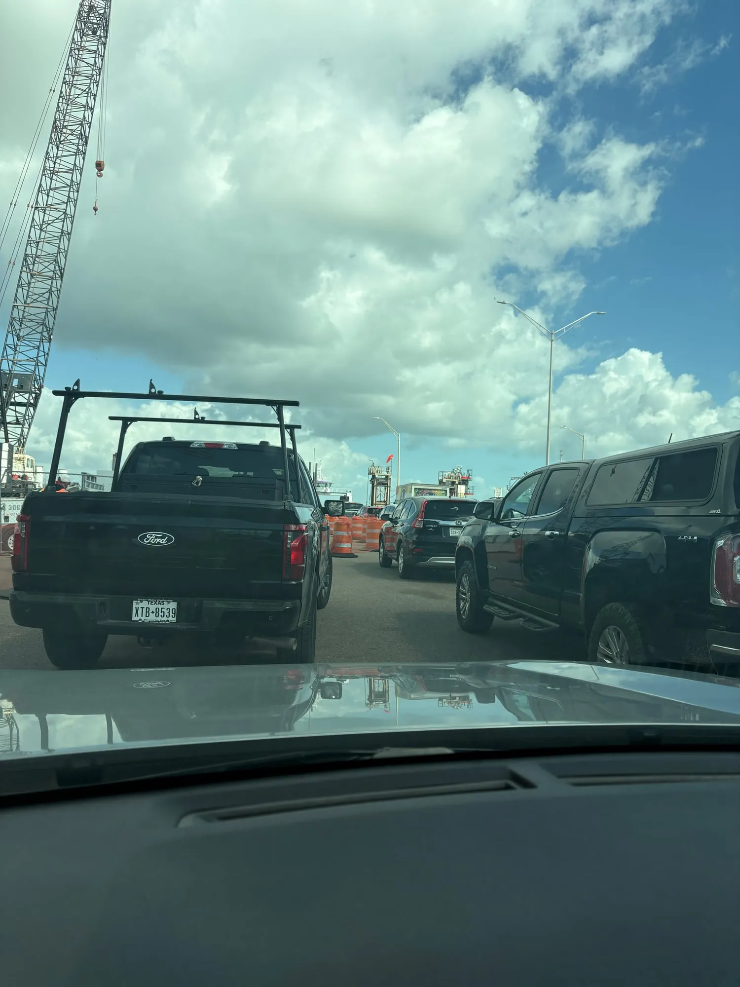 Stopped in a ferry queue behind a black pickup with a ladder rack, orange barrels and a crane ahead under heavy clouds
