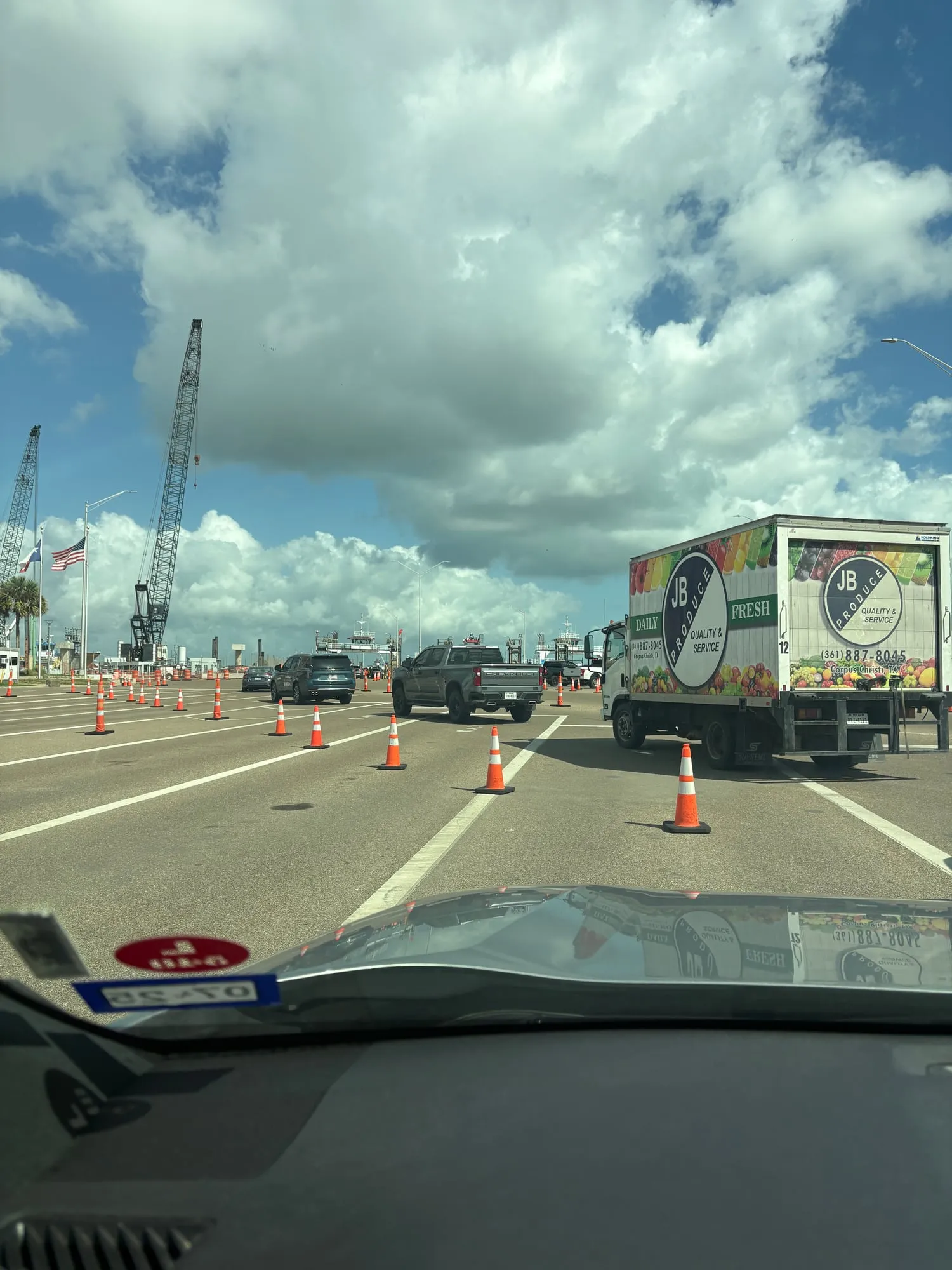 Ferry staging lanes marked off with orange cones, a produce delivery truck and a pickup ahead, construction cranes and flags on the left