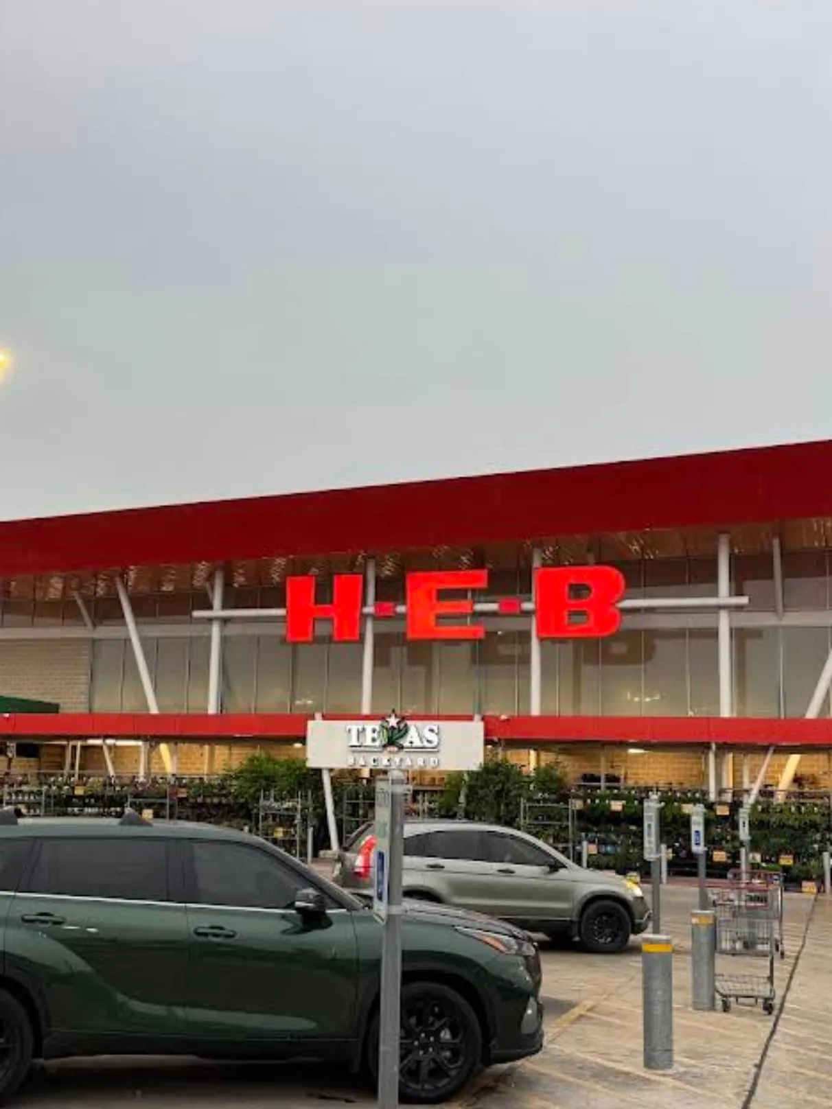 H-E-B storefront under a grey sky, red canopy and big red letters over a glass wall, a green SUV parked in front and the Texas Backyard garden center to the side