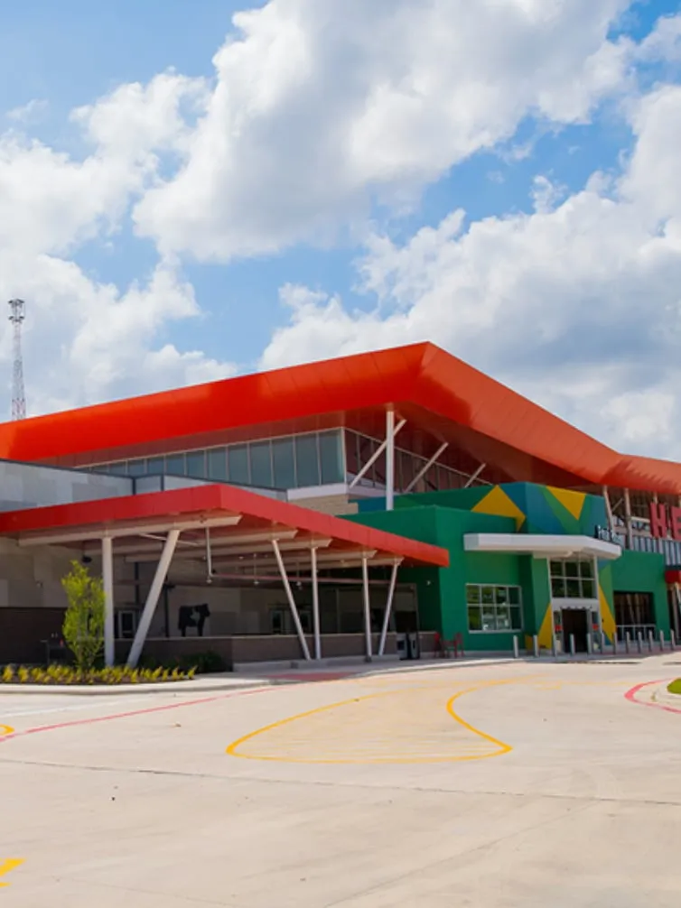 Modern H-E-B with a sweeping orange roofline and a green and yellow geometric entrance under a bright blue sky with puffy clouds