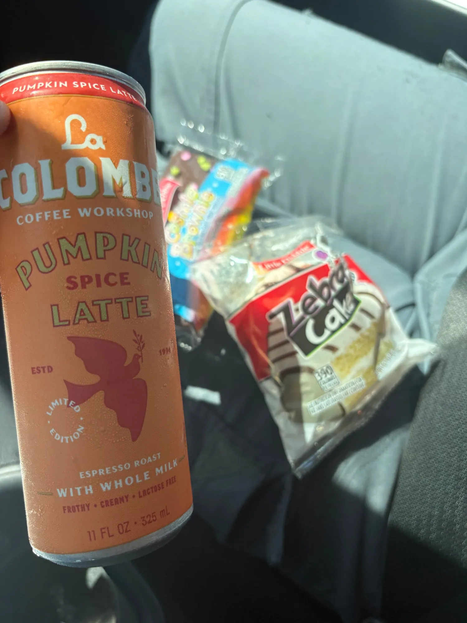 Hand holding an orange can of La Colombe pumpkin spice latte in a car, with a Zebra Cake and a Cosmic Brownie on the seat behind it